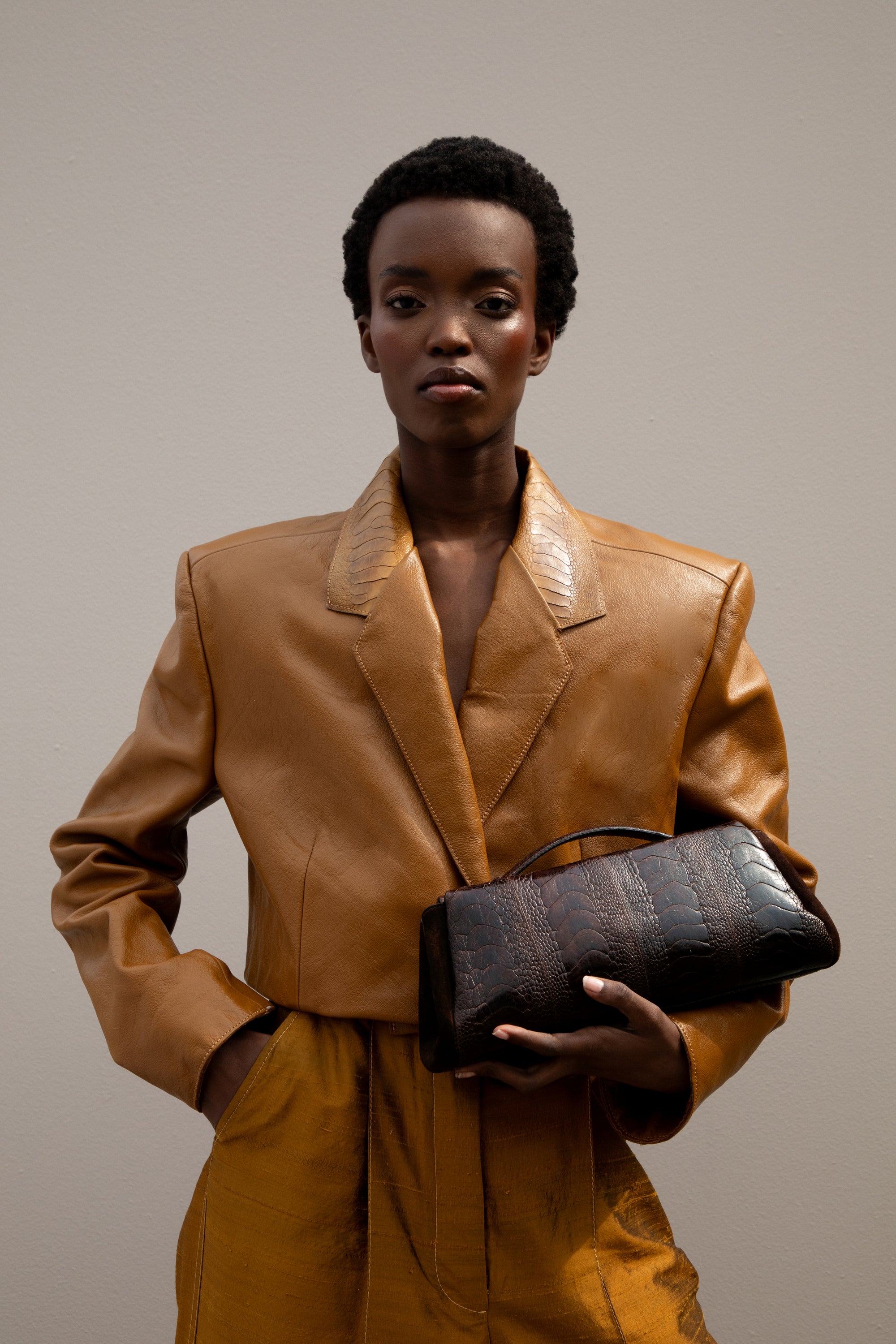 Woman in a brown leather outfit holding a brown clutch bag against a neutral background