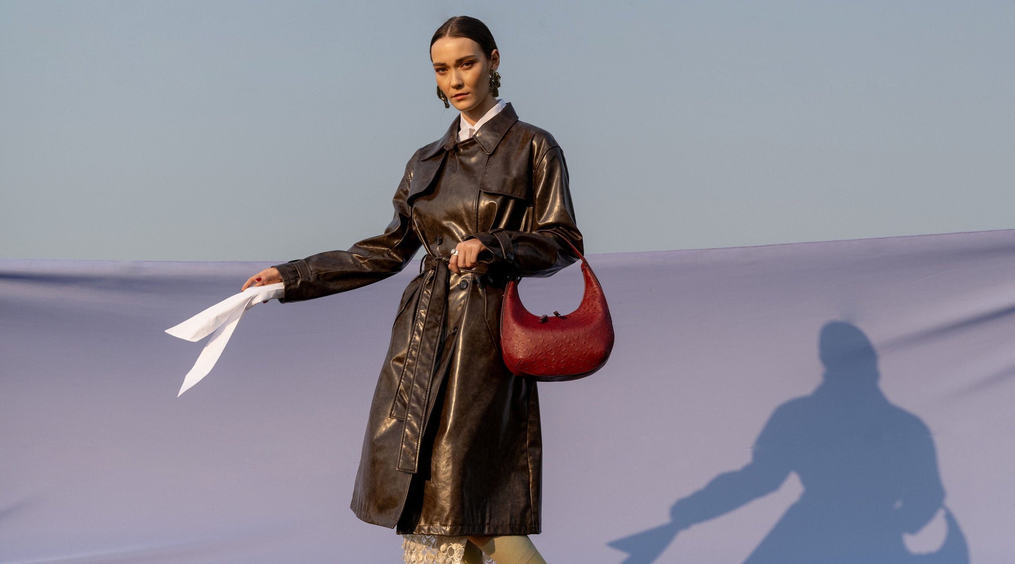 Woman in a black coat holding a maroon ostrich-textured handbag against a clear sky