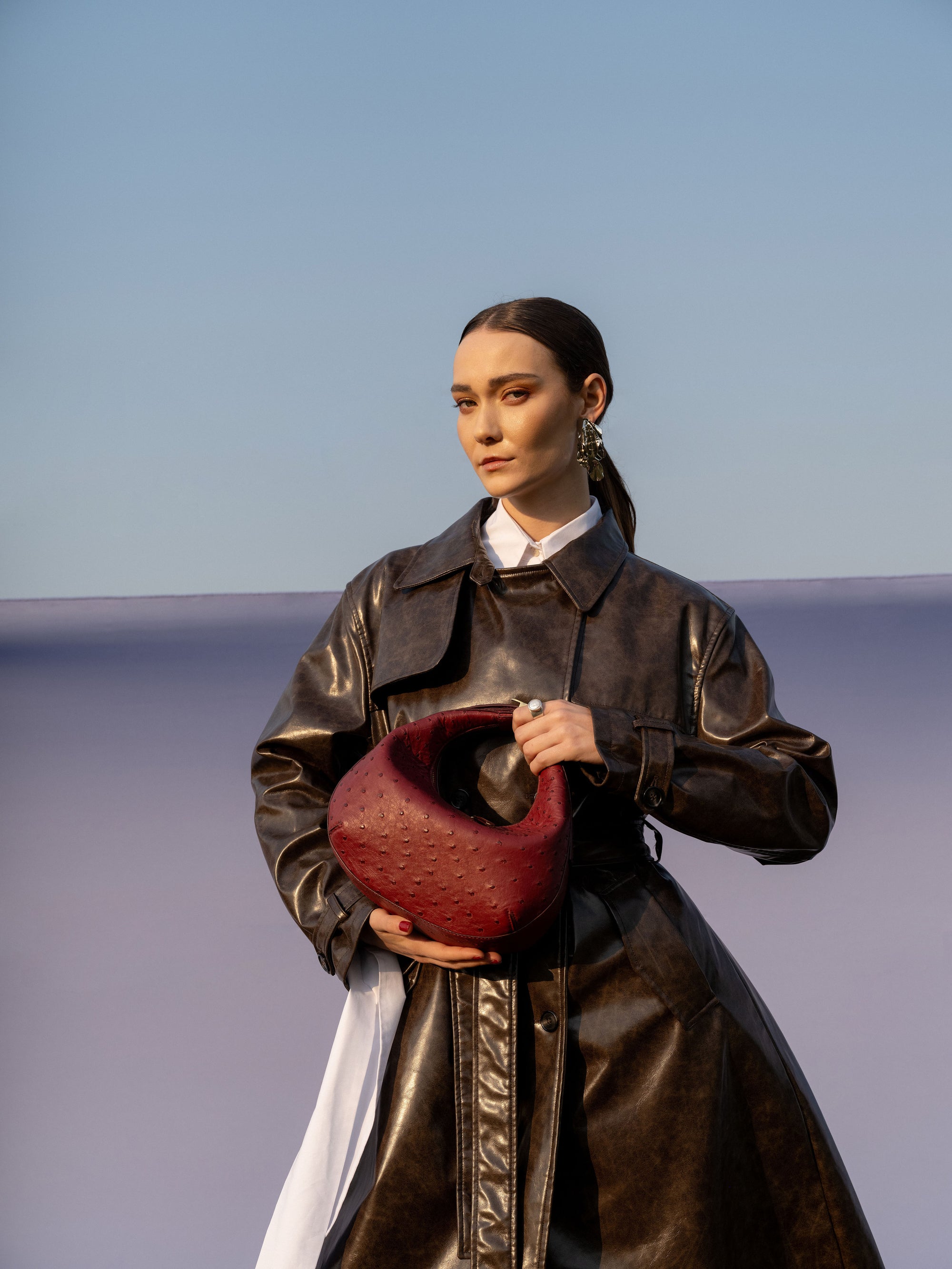Person wearing a long dark coat holding a maroon handbag against a blue sky.