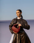 Person wearing a long dark coat holding a maroon handbag against a blue sky.