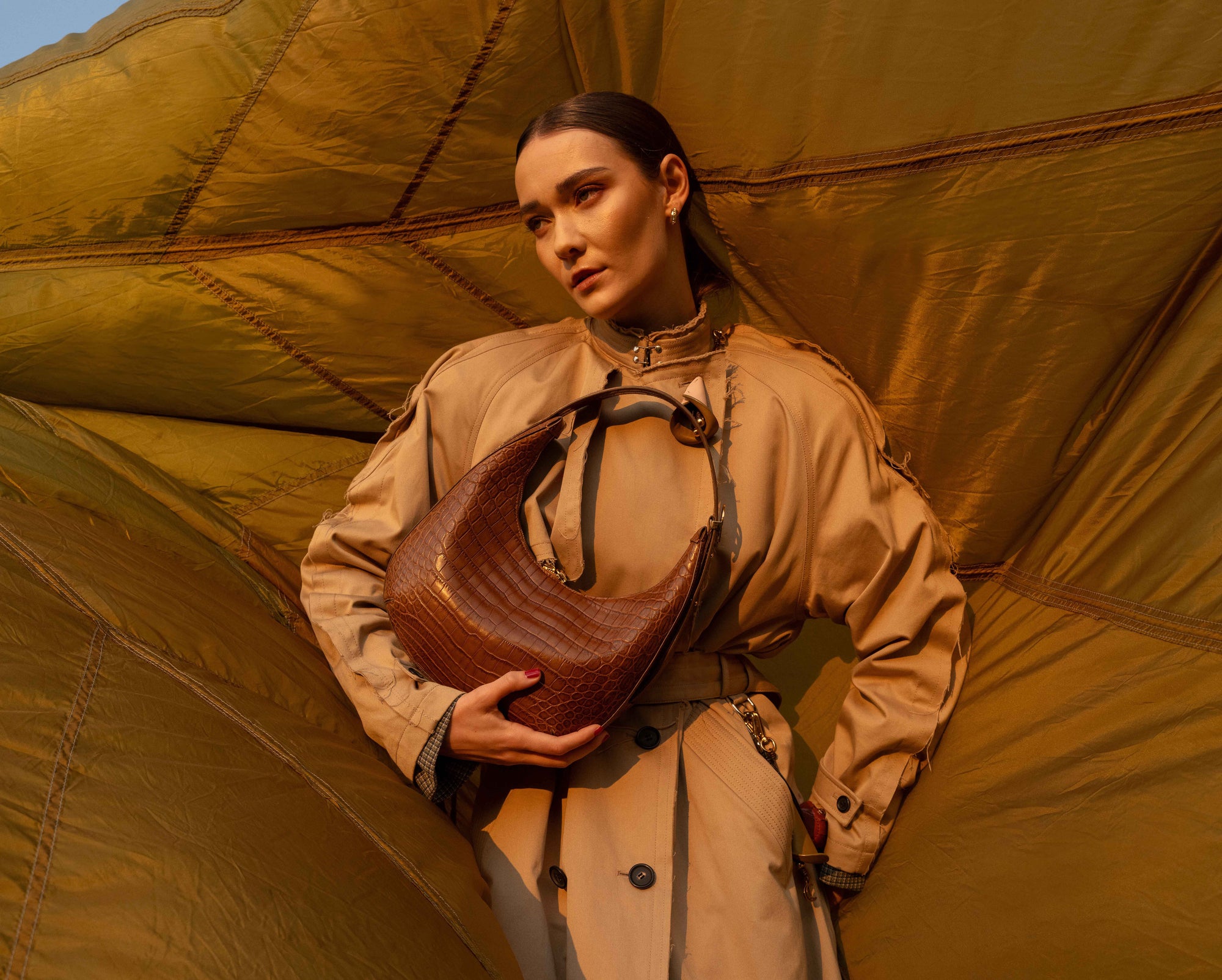 Person in a beige trench coat holding a brown leather bag against a large green leaf-like backdrop.