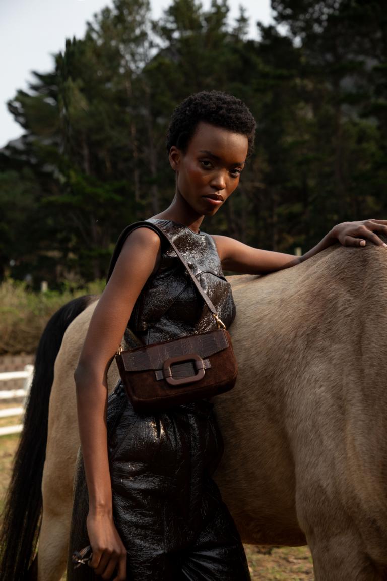 Woman standing next to a horse in an outdoor setting wearing a brown handbag