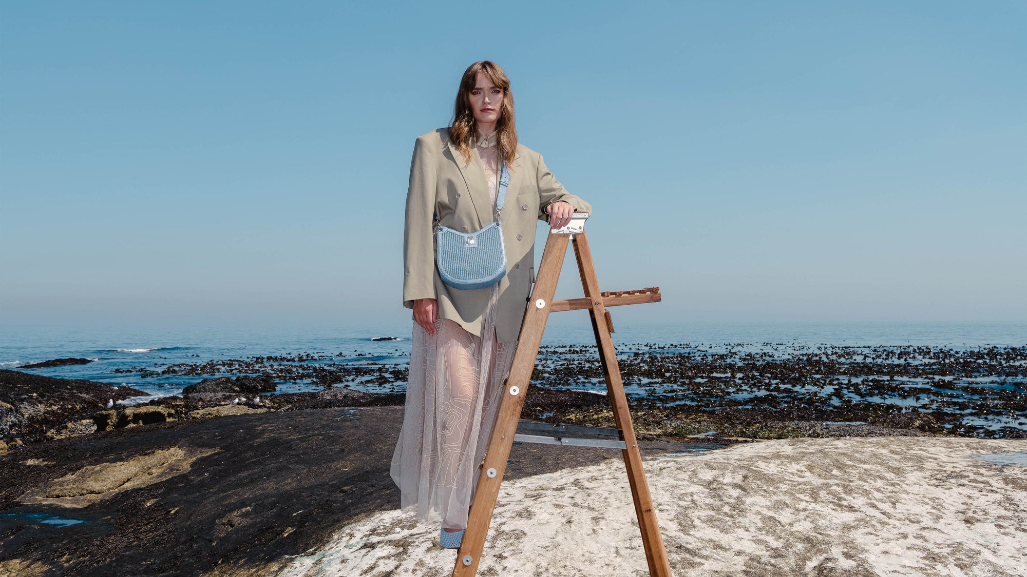 Woman standing on a rocky beach with a wooden ladder with a blue shoulder bag.