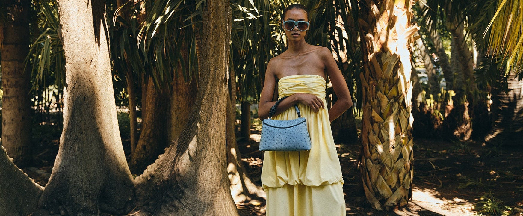 Woman in a yellow dress holding a blue rope handle ostrich leather handbag in a tropical setting with palm trees.