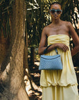 Woman in a yellow dress holding a blue rope handle ostrich leather handbag in a tropical setting with palm trees.
