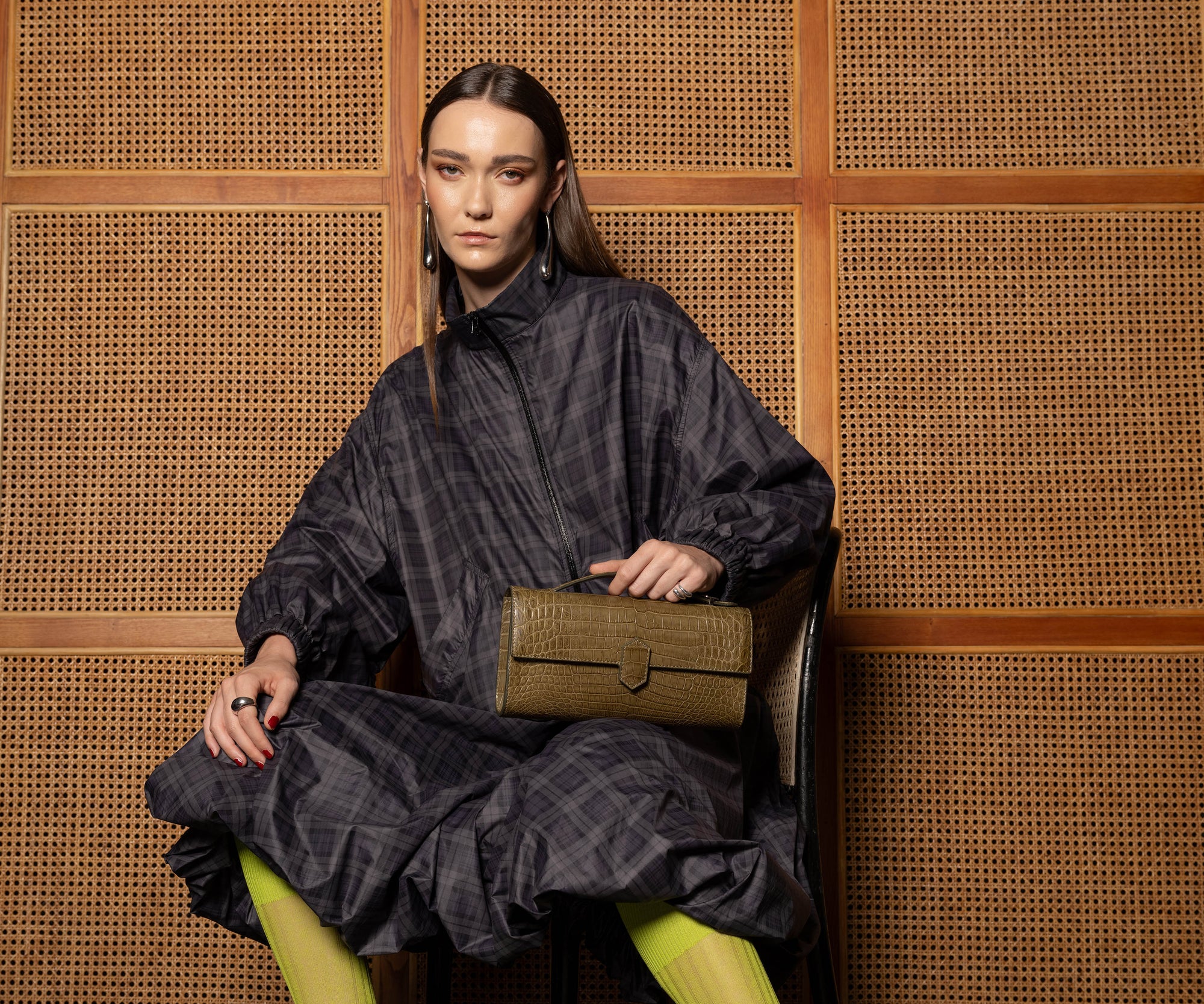 Woman in a dark outfit holding a forest green crocodile-textured clutch handbag against a woven wooden background