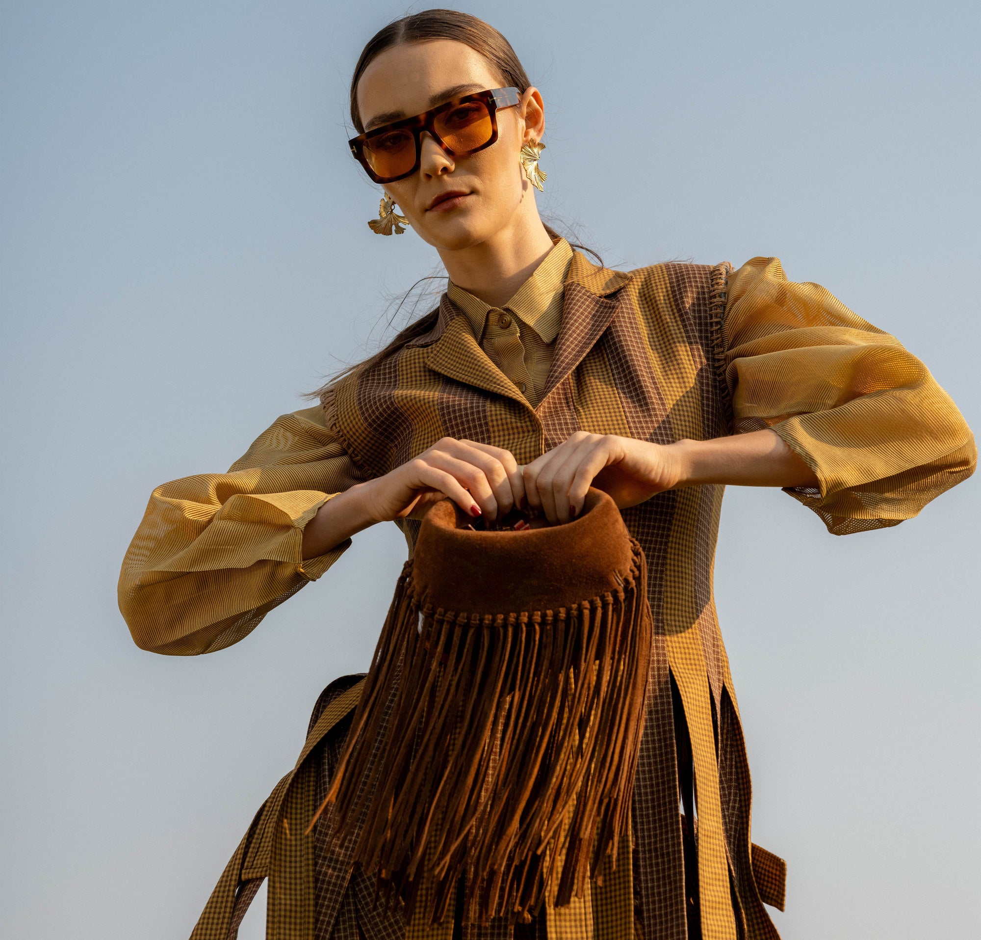 Woman modeling with a brown suede fringed handbag against a clear sky.