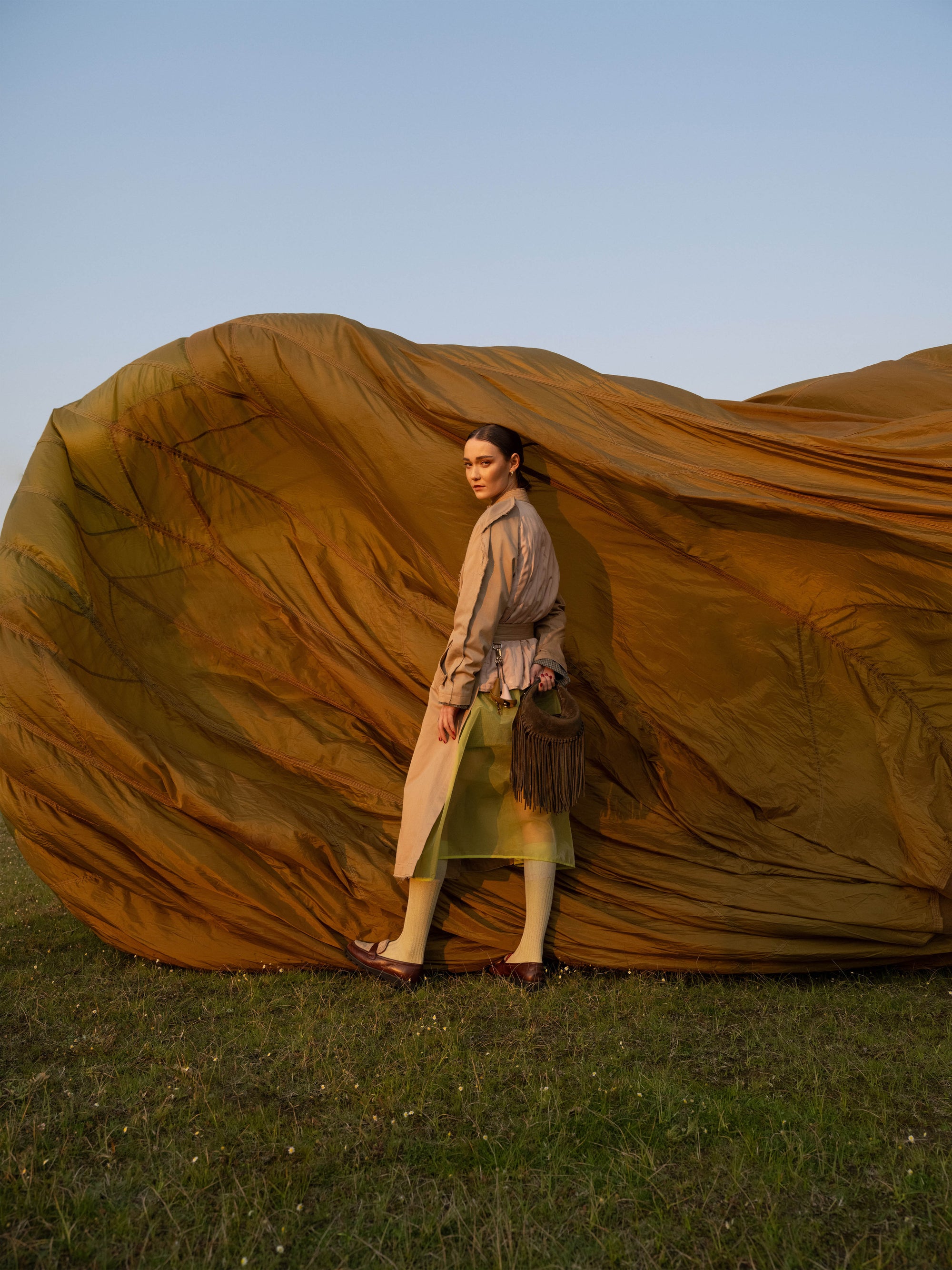Woman standing infront a large brown inflatable object, wearing a beige coat and holding a olive suede  handbag with a fringe.