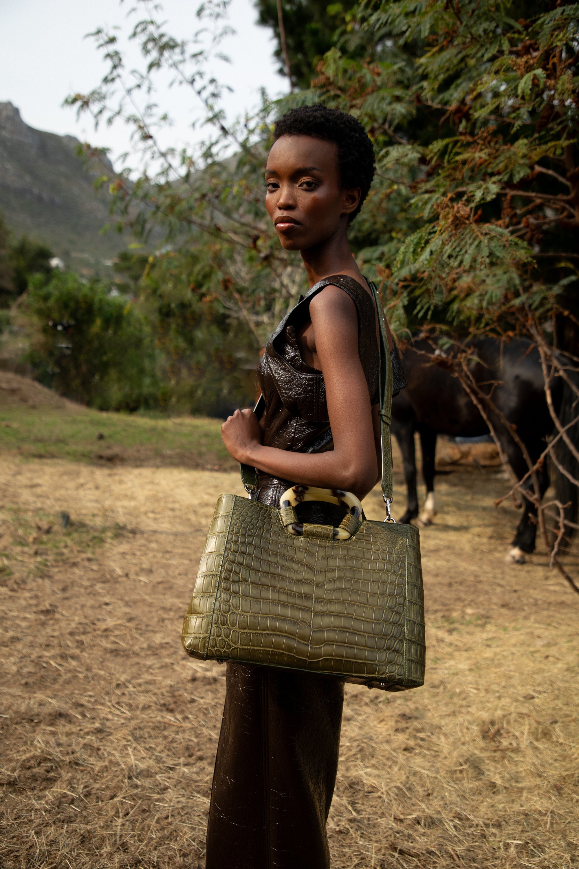 Woman holding a green handbag in a natural setting with trees and mountains in the background