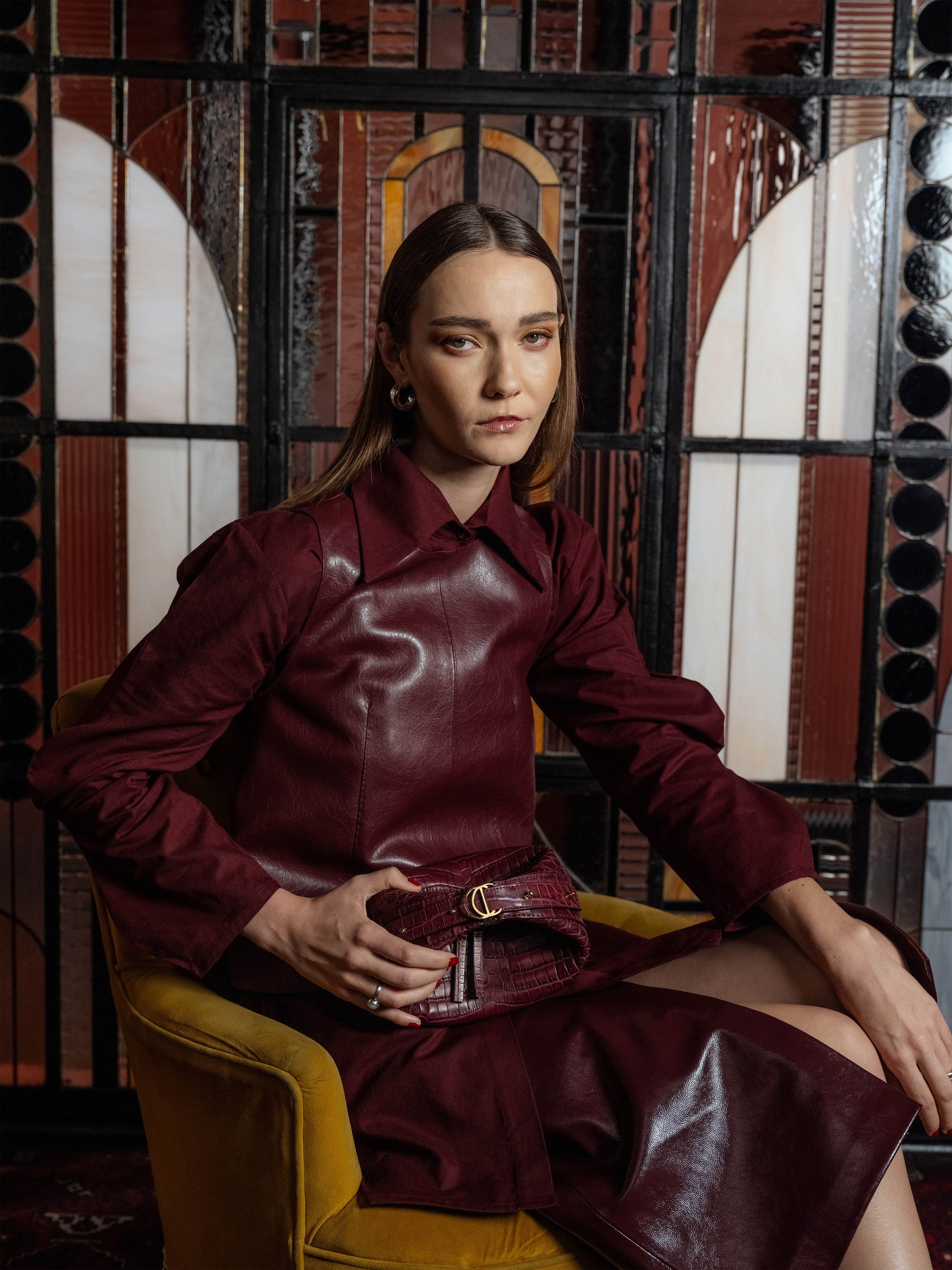 Woman in a burgundy leather outfit sitting on a yellow chair holding a burgundy crocodile leather handbag