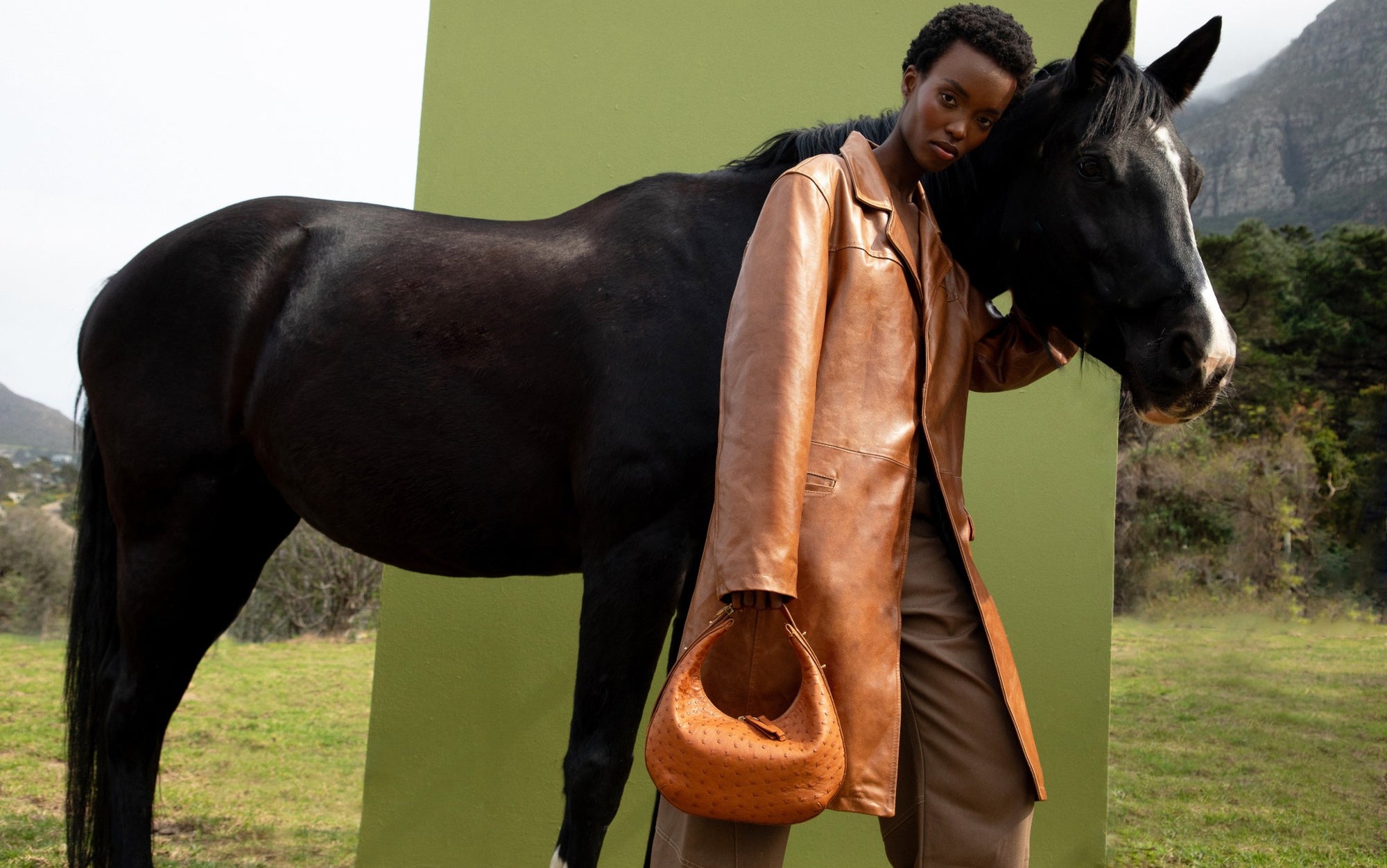 Woman holding a cognac ostrich leather bag next to a horse in a natural setting