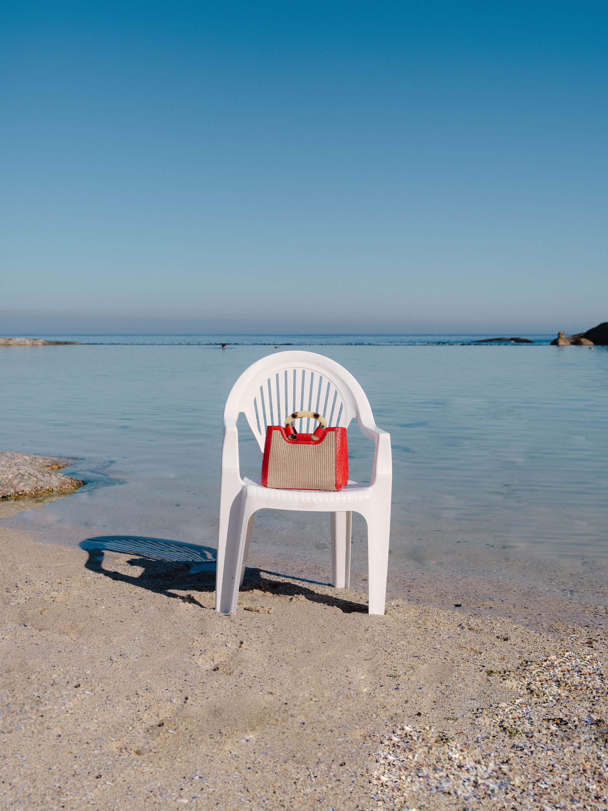 White chair with a red and beige handbag on a sandy beach with clear blue water and sky.