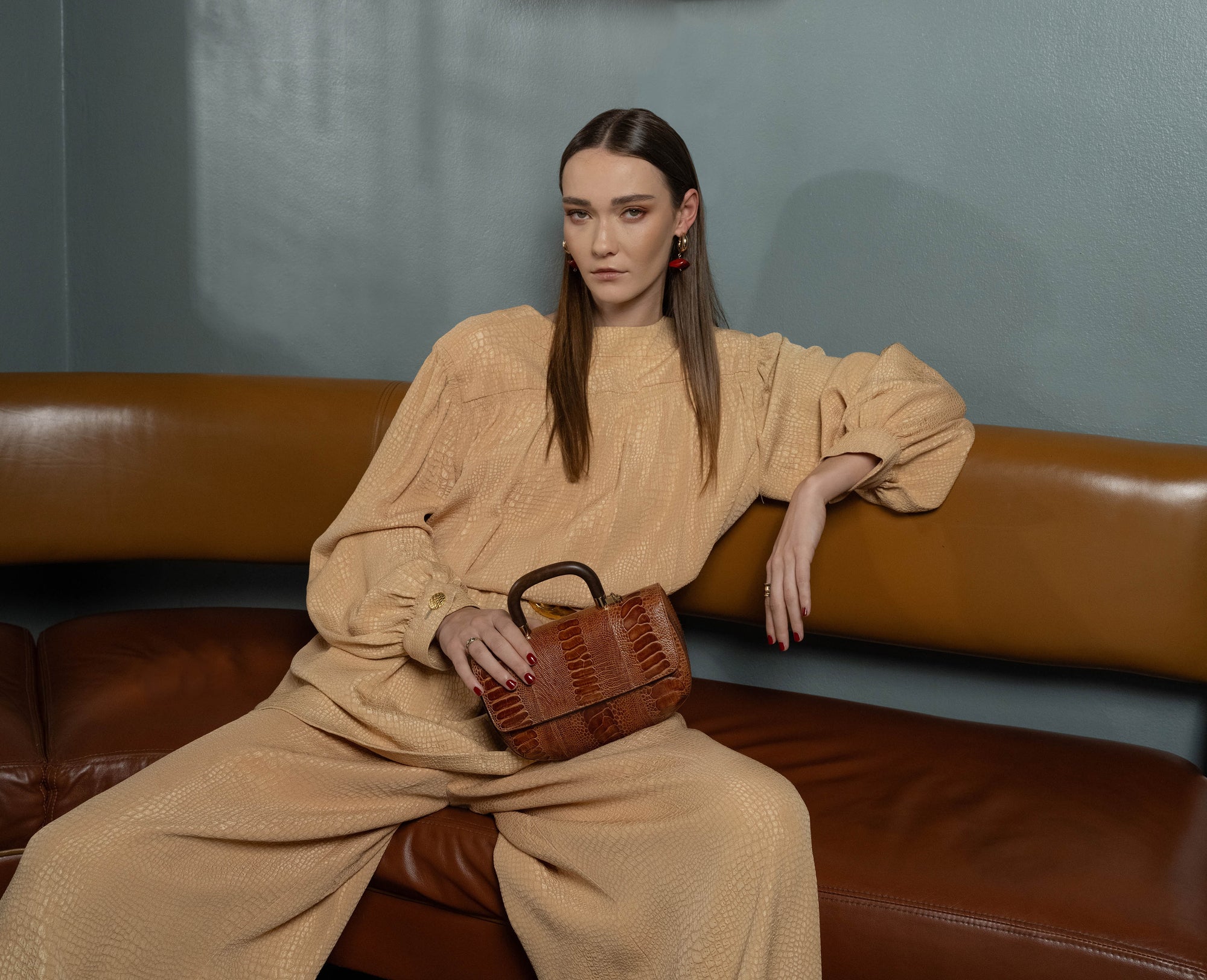 Woman in beige outfit sitting on a brown leather couch holding a brown handbag.