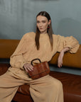 Woman in beige outfit sitting on a brown leather couch holding a brown handbag.